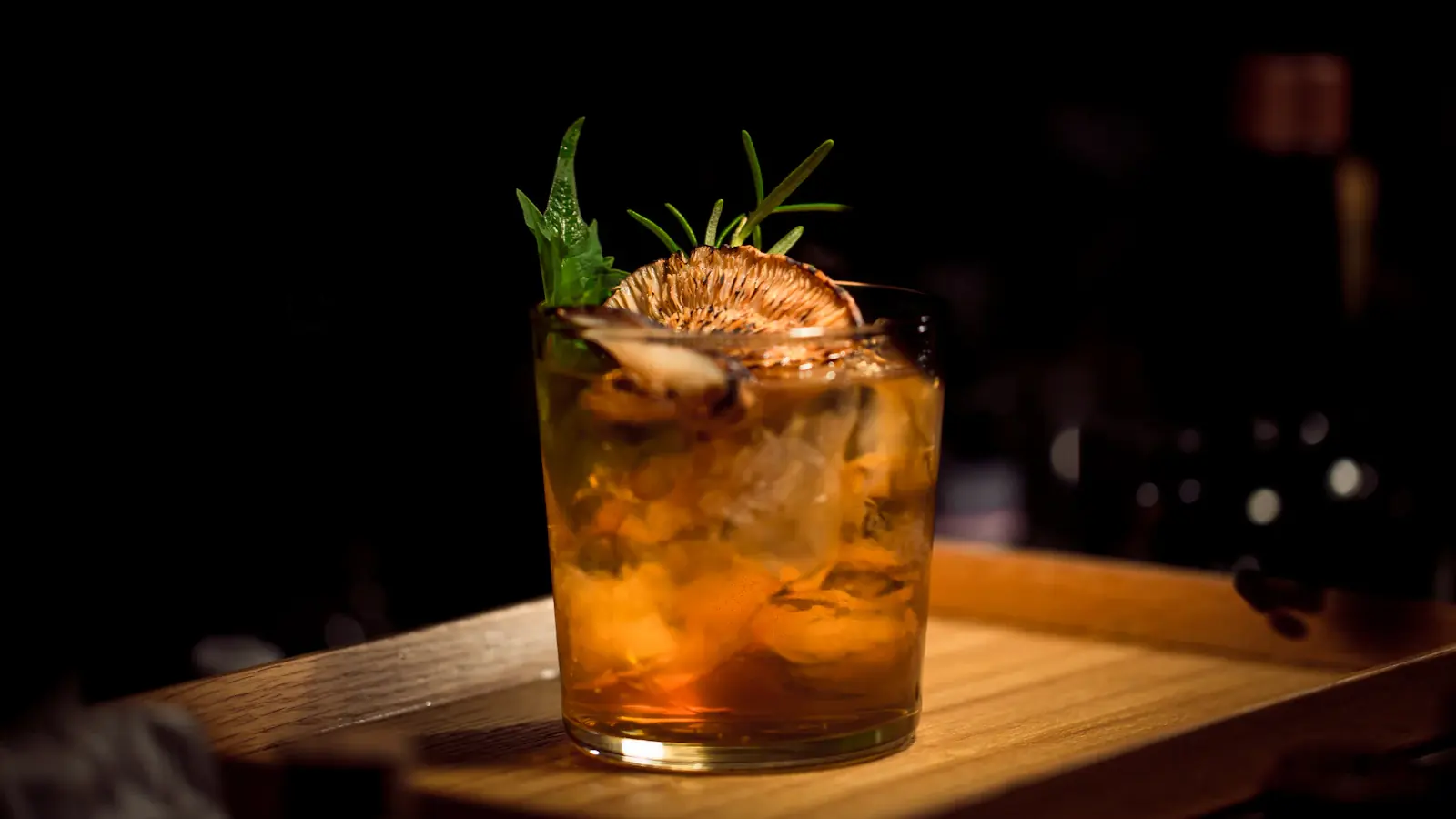 Cóctel con hielo y una ramita de romero sobre una tabla de madera, en la penumbra de un bar