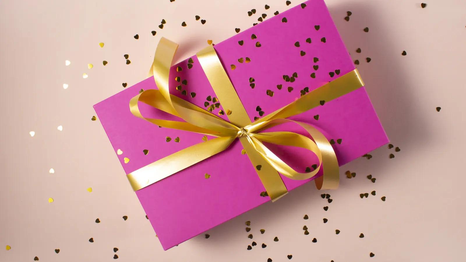 Caja de regalo envuelta en papel fucsia con lazo dorado, rodeada de confeti